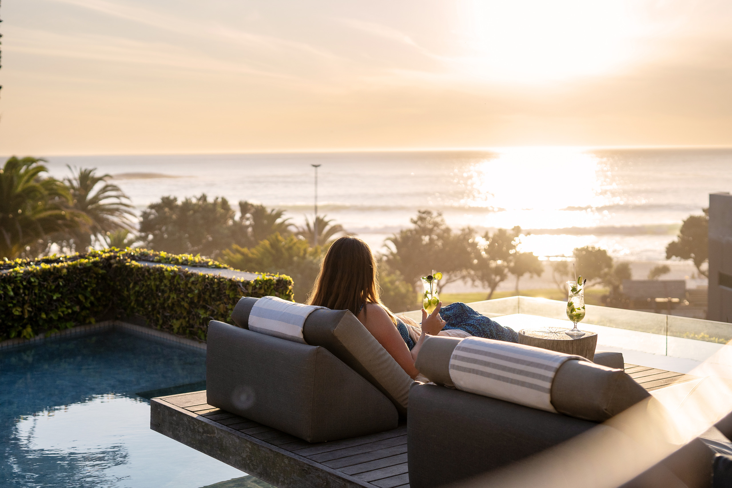 POD-Camps-Bay-view A woman lounges poolside with cocktails, overlooking the ocean from one of the luxury hotels in Cape Town
