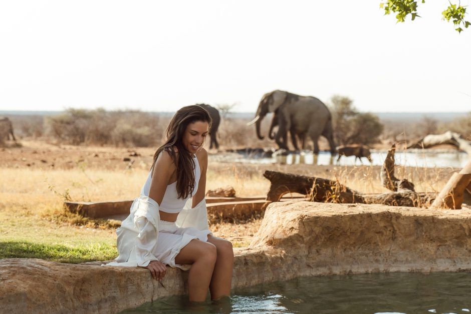 last_word_madikwe_garden_and_pool_1 A woman sits by a pool dipping her feet in the water, with elephants drinking at a nearby watering hole in a dry, open landscape.